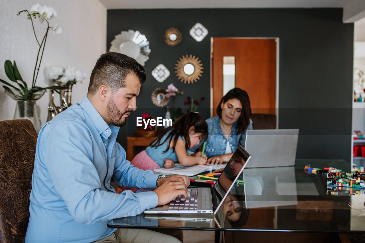 Side view of concentrated young latin american self employed man in shirt working online on laptop sitting at table near wife and little daughter painting in sketchbook at home