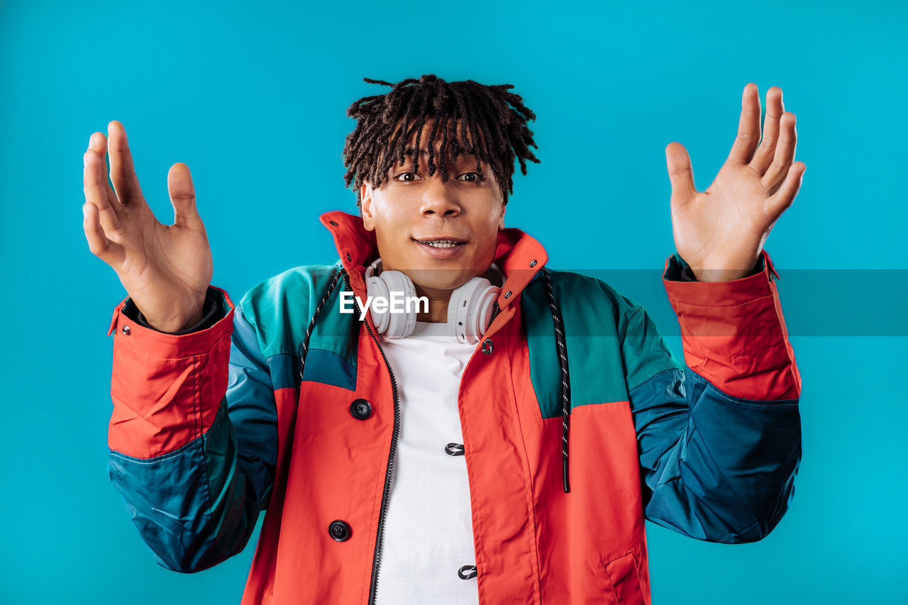 one person, studio shot, portrait, colored background, emotion, clothing, gesturing, men, waist up, person, arm, hand, front view, blue, jacket, happiness, fun, young adult, positive emotion, looking at camera, limb, indoors, child, smiling, human limb, arms raised, adult, blue background, standing, hand sign, cheerful, hand raised, casual clothing, childhood