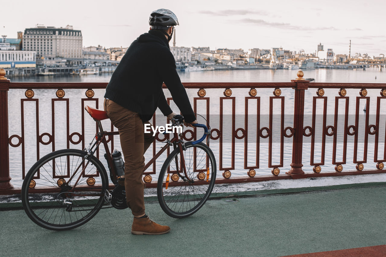 Side view of male cyclist with bicycle standing on bridge over river against sky in city
