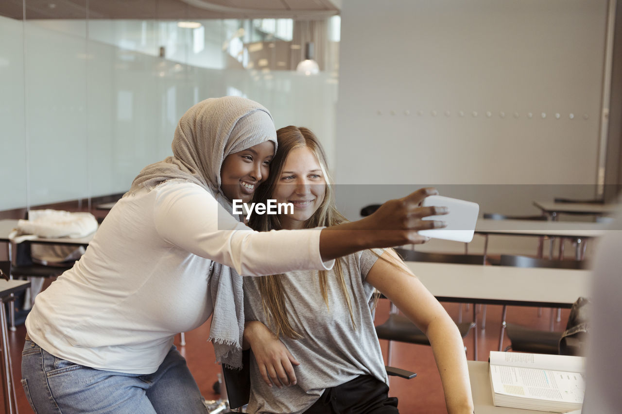 Smiling woman in hijab taking selfie with friend on smart phone at classroom