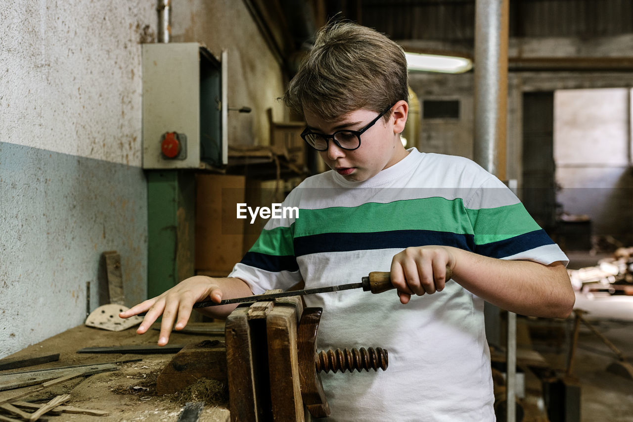 Serious teenage boy grinding piece of wood in vice using metal rasp while working in shabby workshop