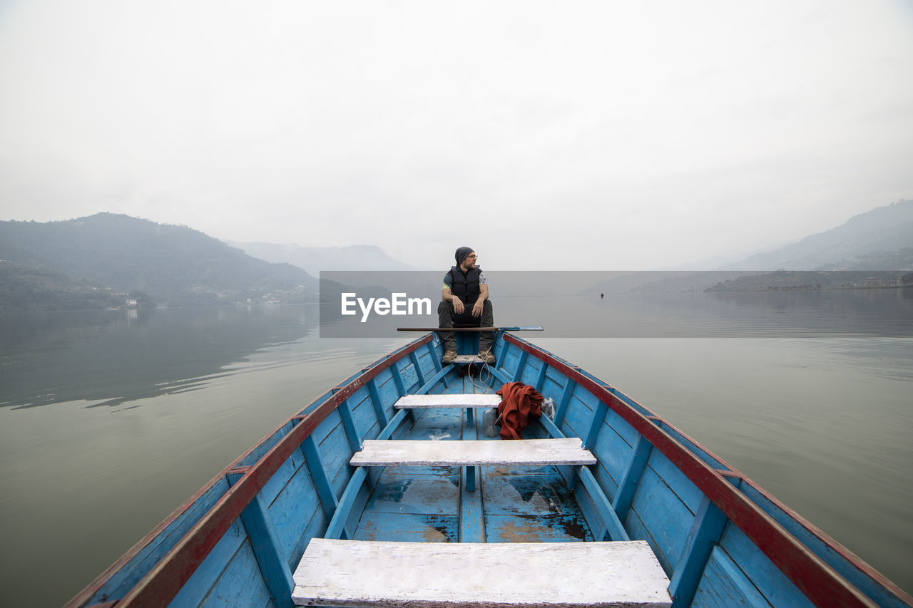 Male explorer floating in blue wooden boat on calm lake in misty morning during vacation in nepal