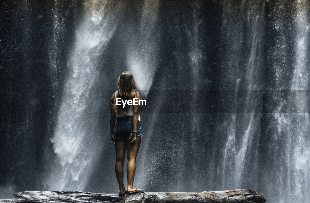 REAR VIEW OF WOMAN STANDING ON ROCK AT WATERFALL