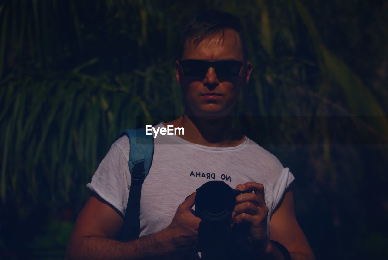 PORTRAIT OF YOUNG MAN WEARING SUNGLASSES WHILE HOLDING CAMERA