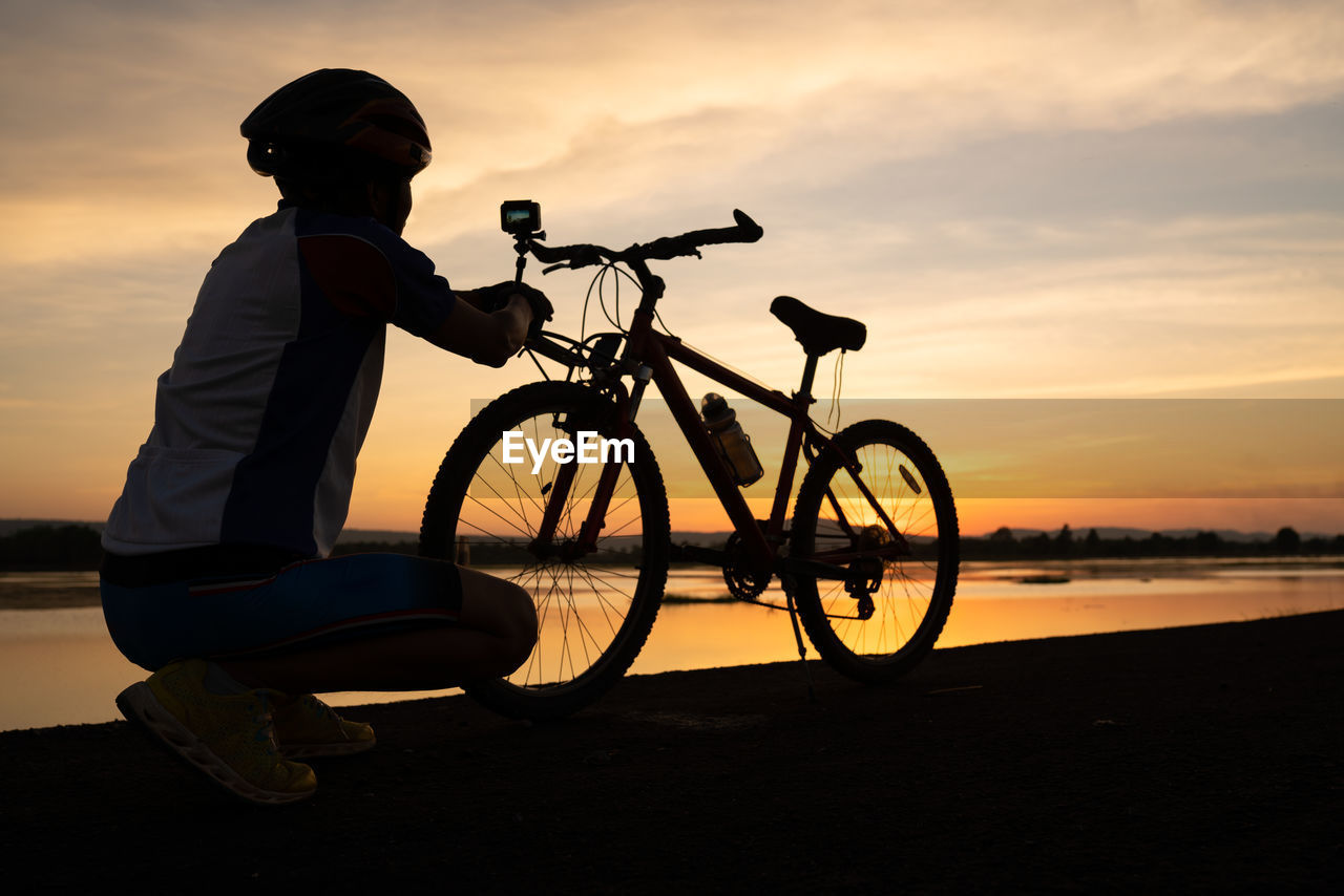 Side view of man crouching by bicycle at lakeshore during sunset