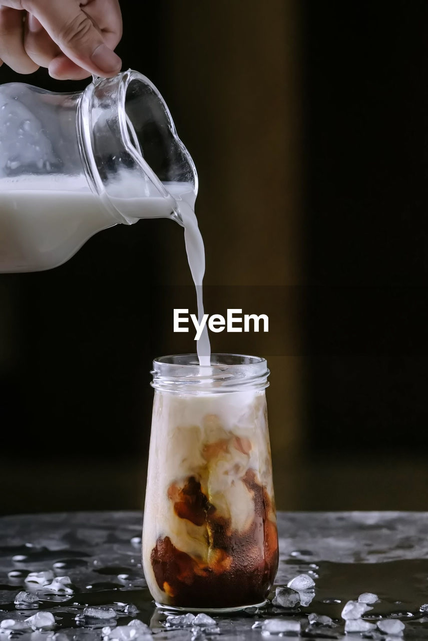 Cropped hand pouring milk in iced coffee on table