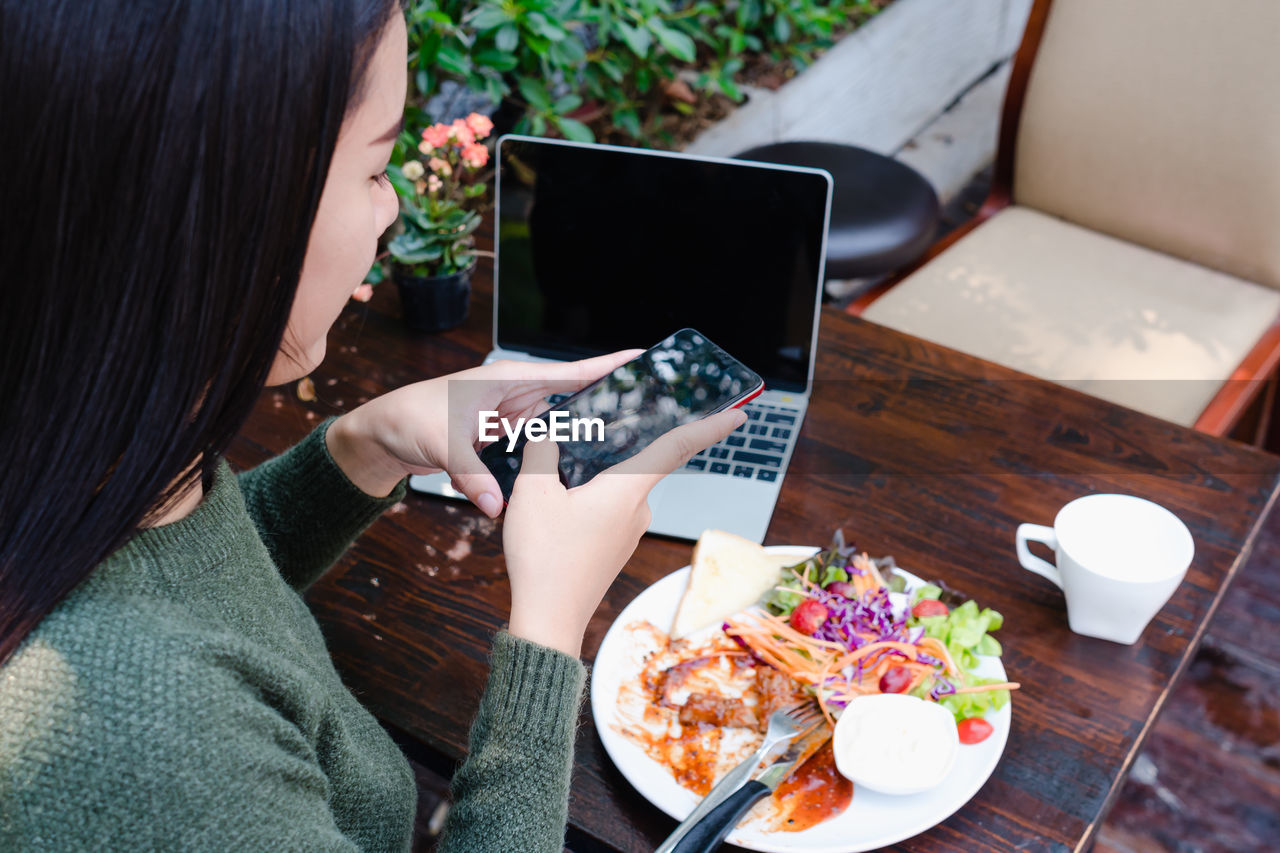 High angle view of woman photographing food with smart phone by laptop on table