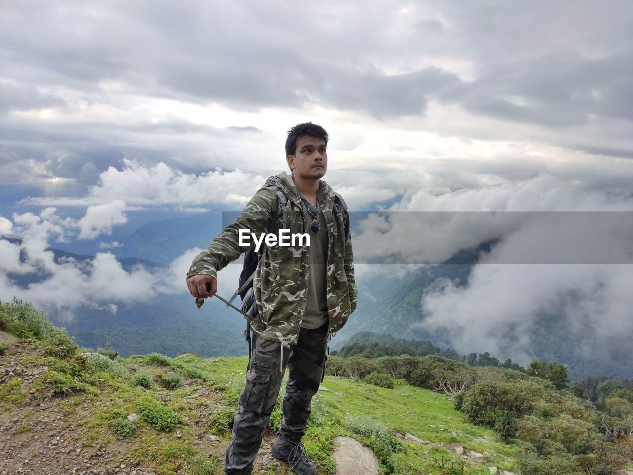 Young man standing by cliff against cloudscape