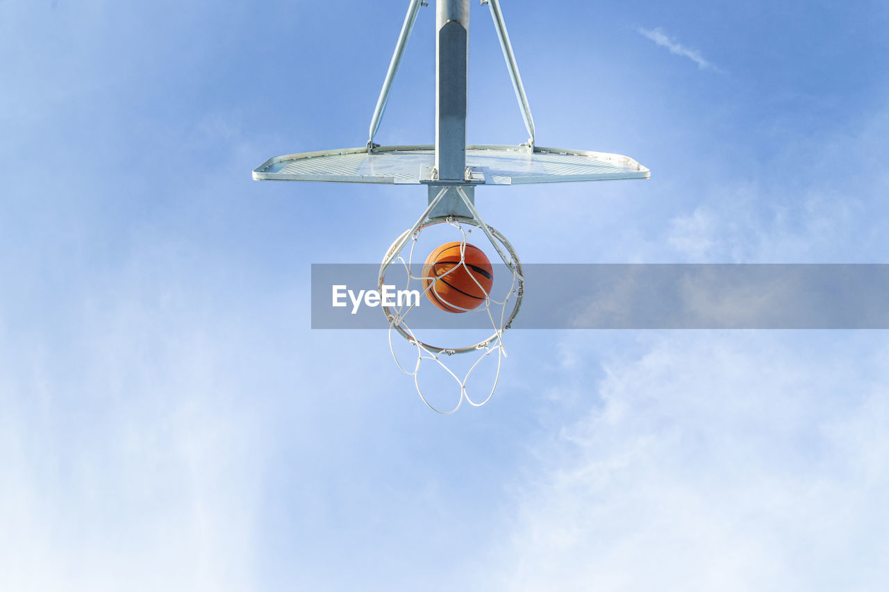 From below of orange basketball falling into round hoop with net placed against blue sky on sunny summer day in city