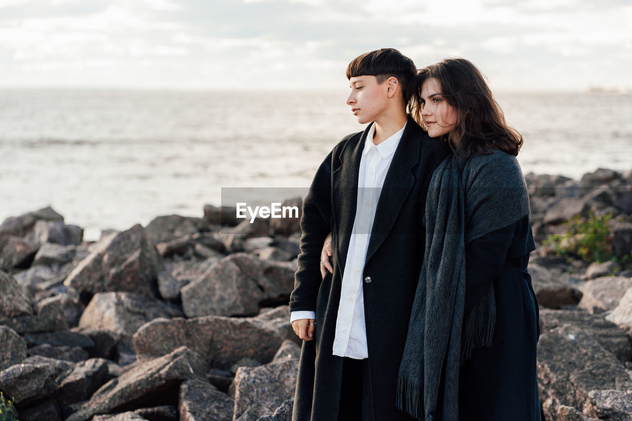 Lesbian women embracing while standing on rock against sea and sky