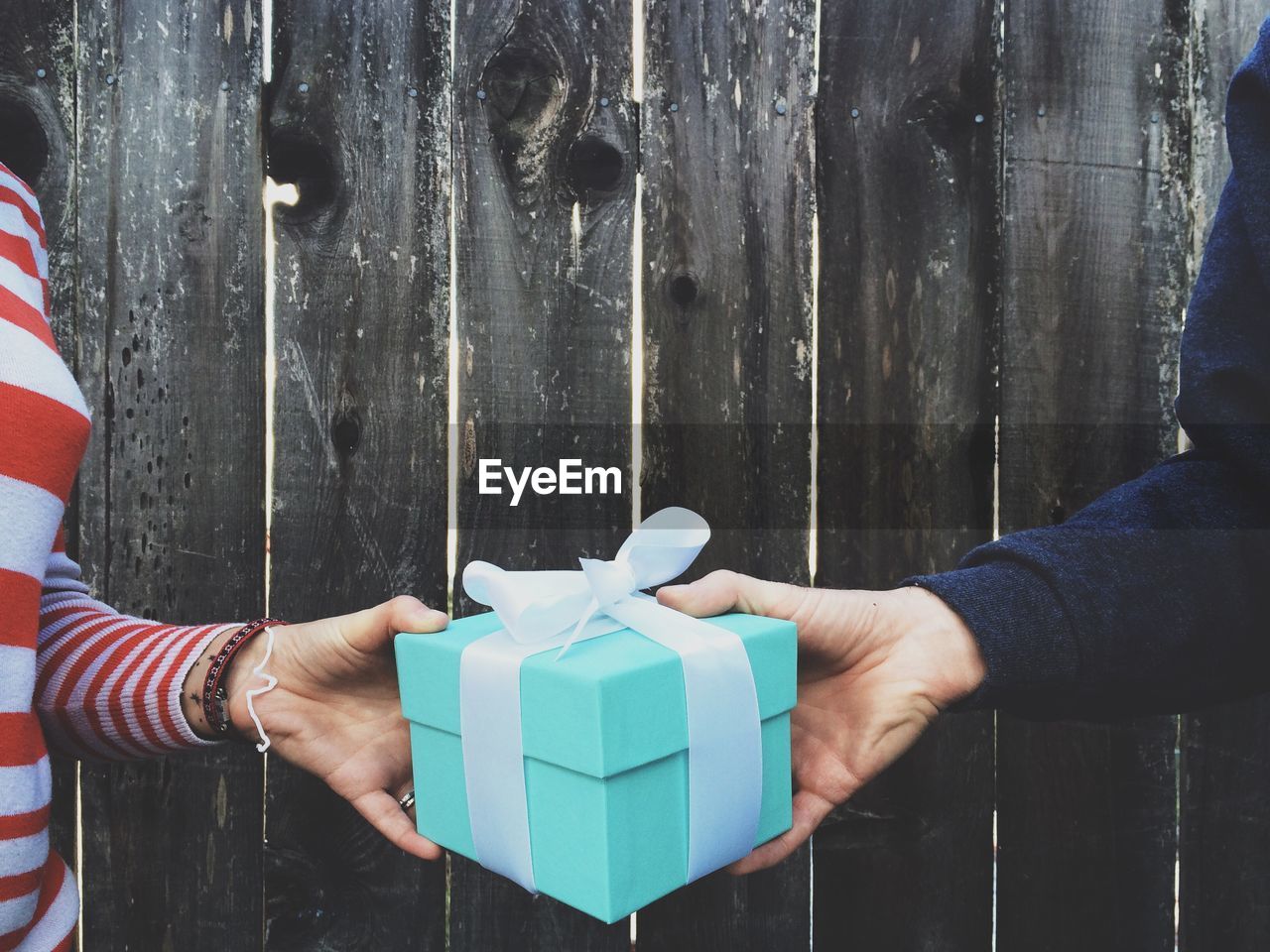 Cropped image of hands holding gift box against wooden fence