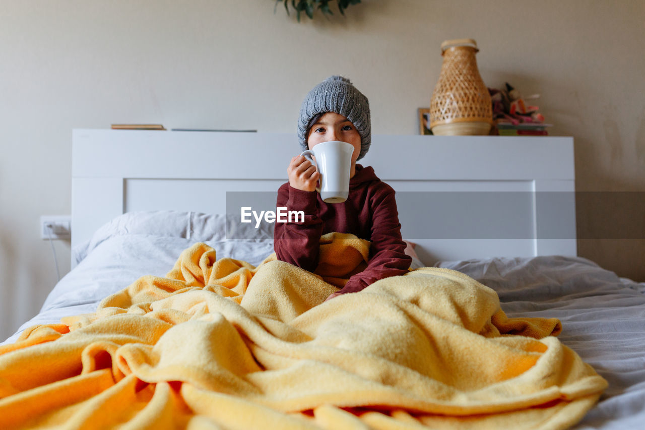 Girl wearing grey warm hat sits on the bed under warm blanket