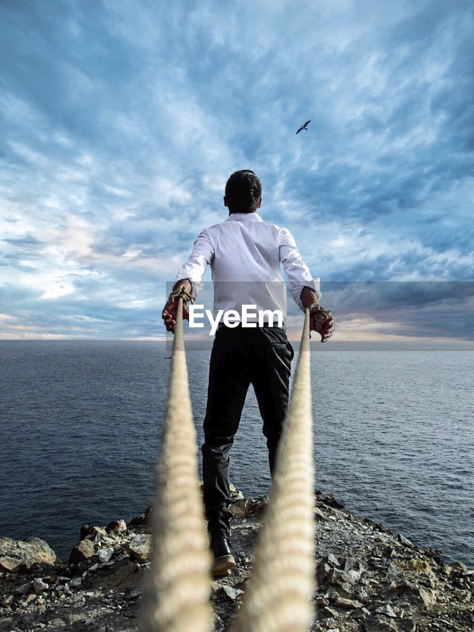 Rear view of man holding ropes while standing at beach against cloudy sky