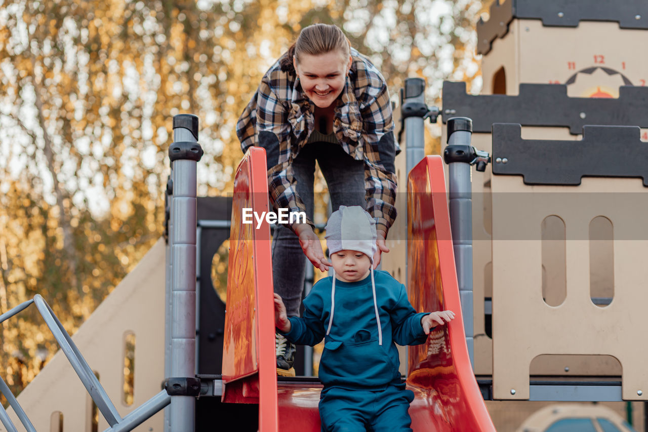 A boy, person with down syndrome walks in the park with his mother, going down the children's slide