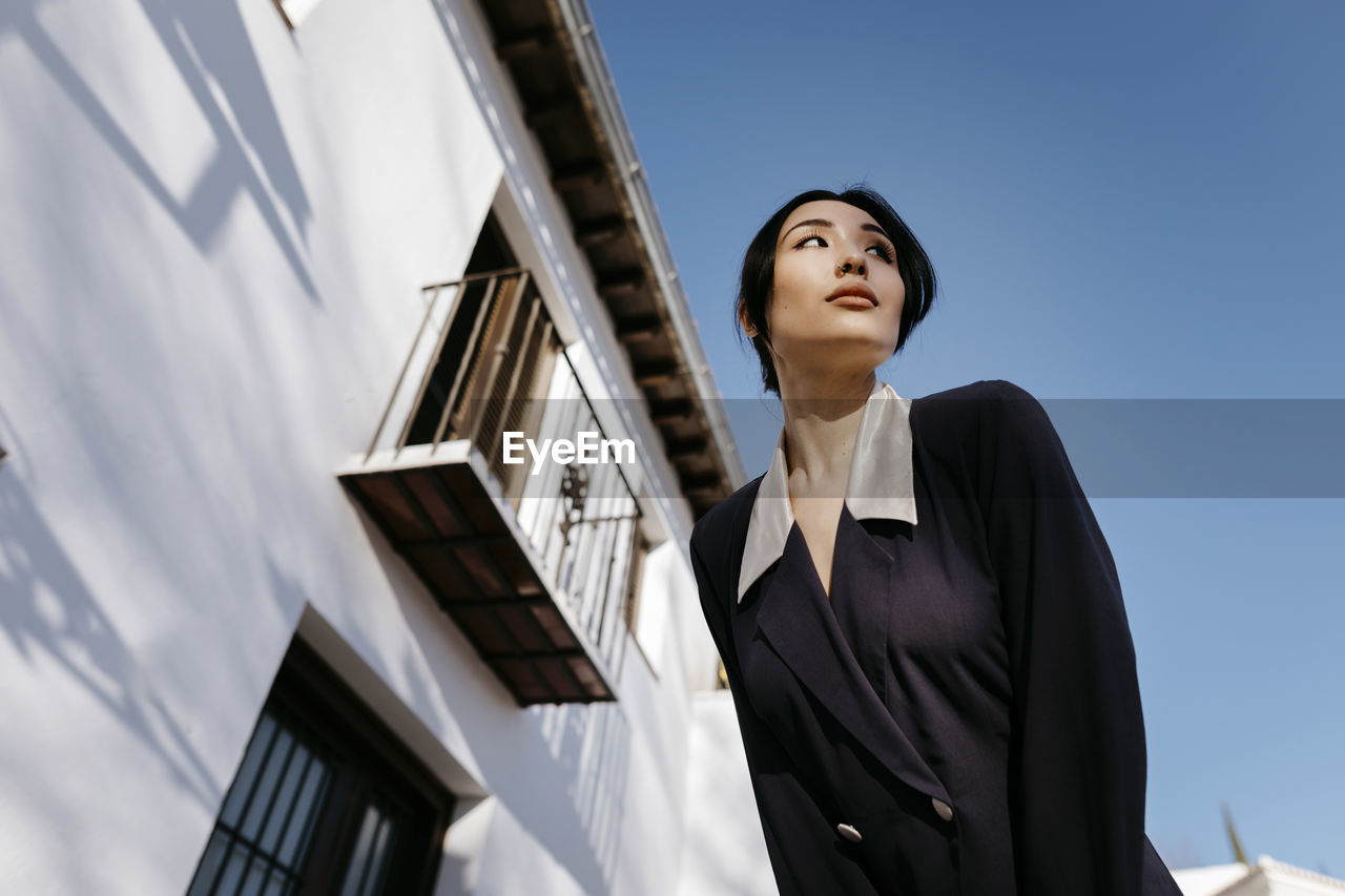 Beautiful woman looking away while standing by building during sunny day