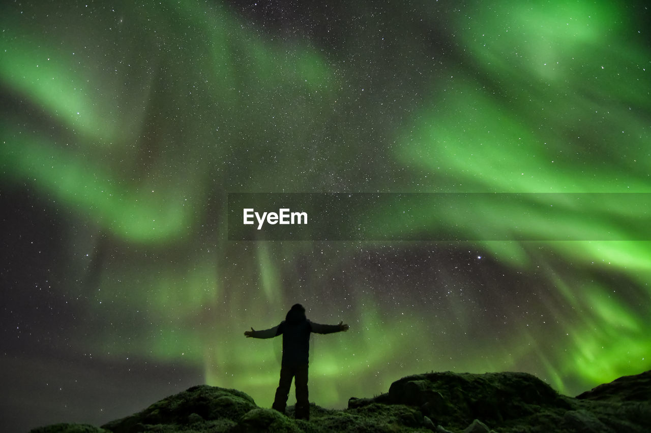 Low angle view of silhouette mature man with arms outstretched standing against northern lights at night