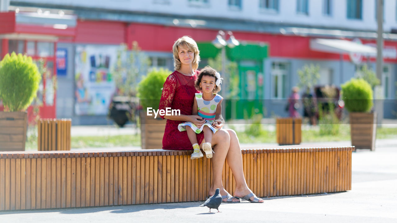 Portrait of mother with cute daughter sitting on seat in city