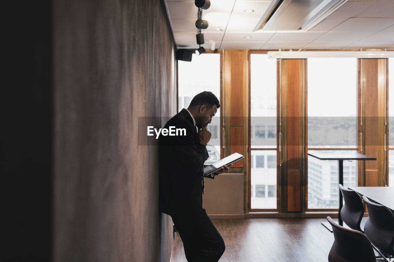 Side view of male entrepreneur with hand on chin holding digital tablet leaning against wall in board room at office