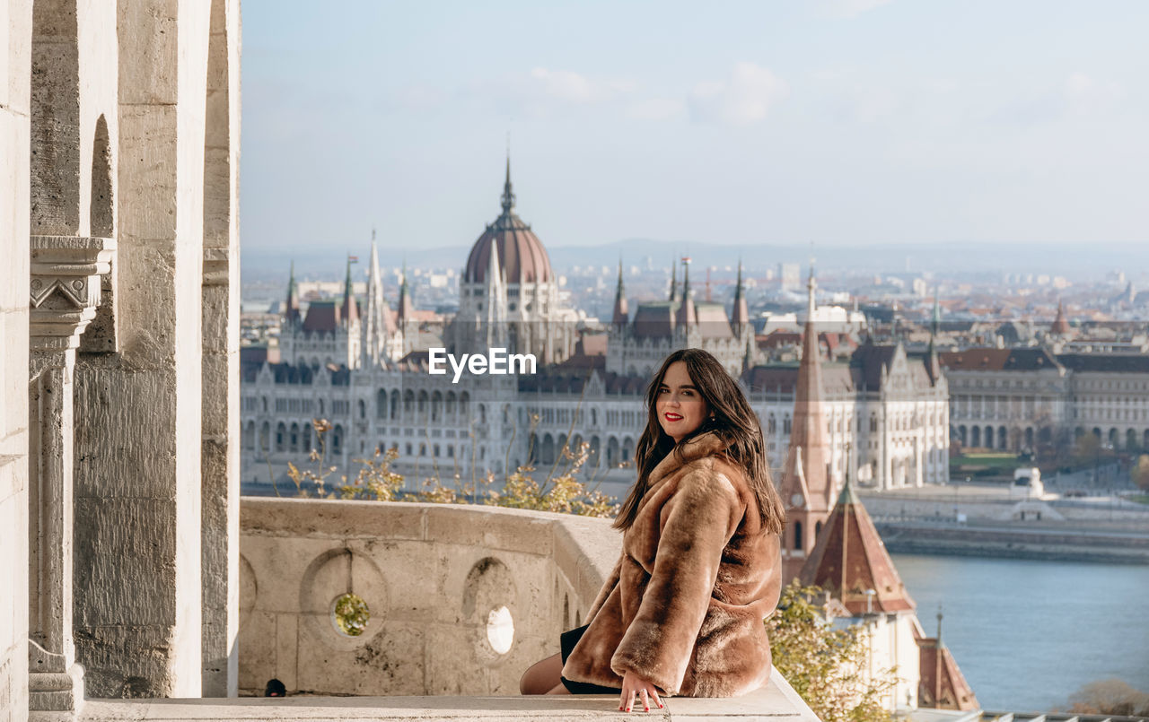 Portrait of beautiful young woman on balcony overlooking hungarian parliament in budapest, hungary