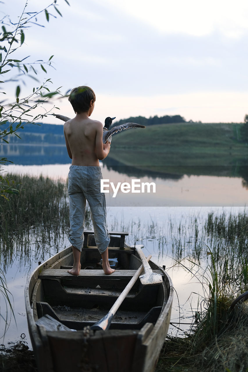 Rear view of boy standing on boat against sky with flying bird