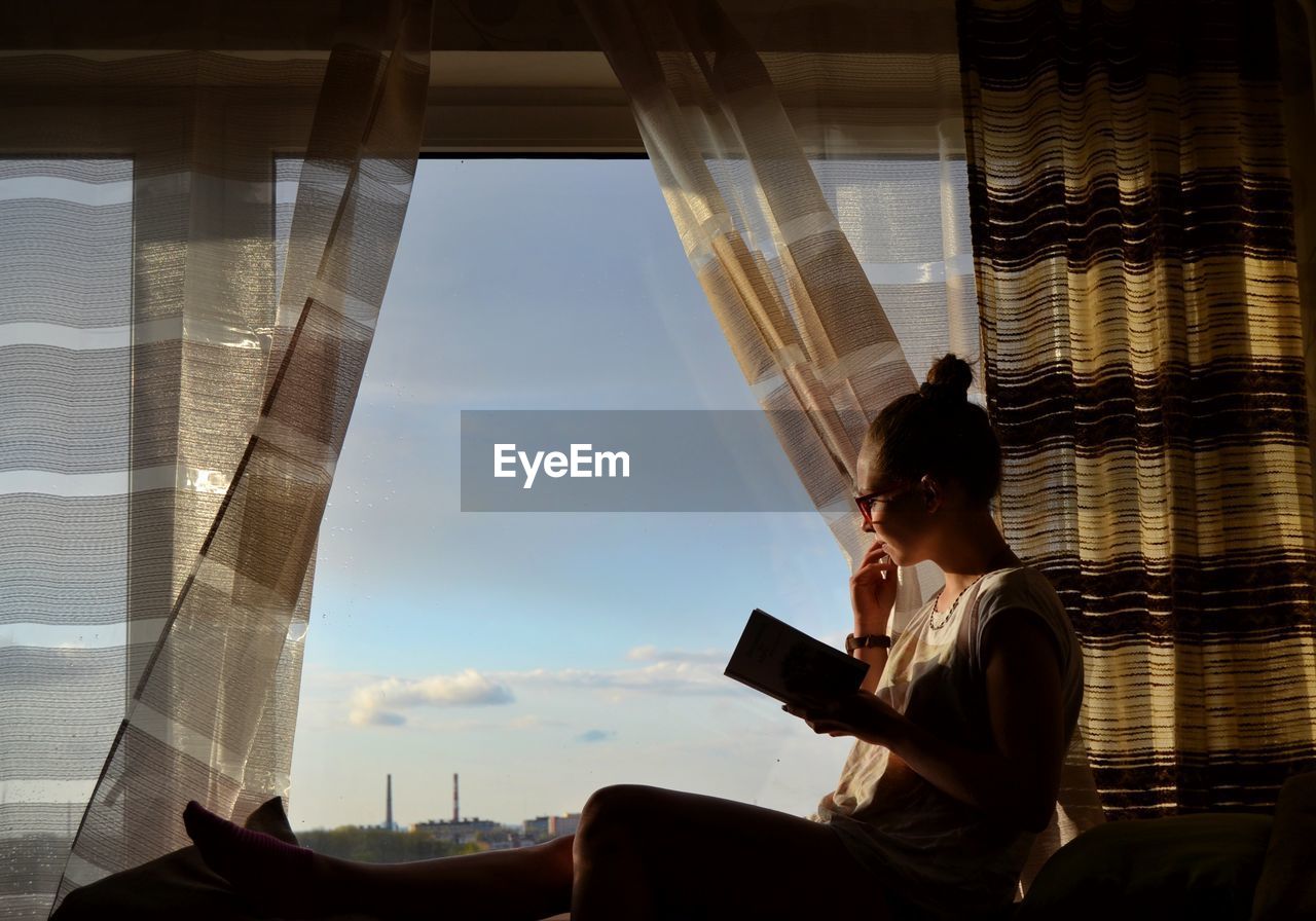 Side view of woman with book sitting on window at home