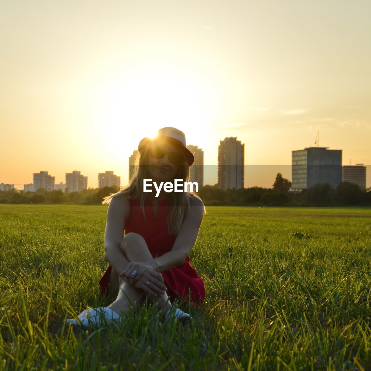 Mid adult woman sitting on grassy field against city during sunset