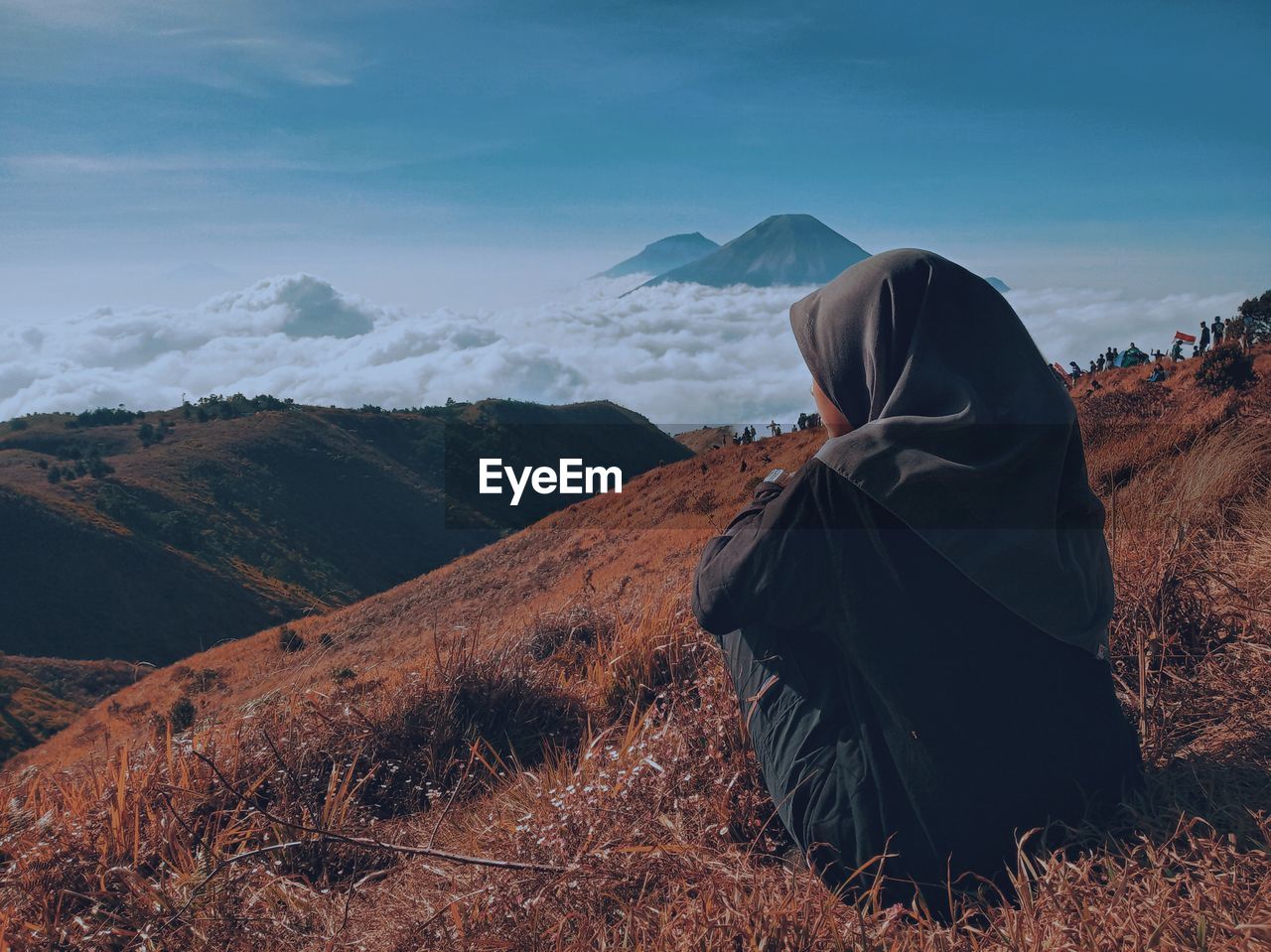 REAR VIEW OF PERSON SITTING ON MOUNTAIN AGAINST SKY