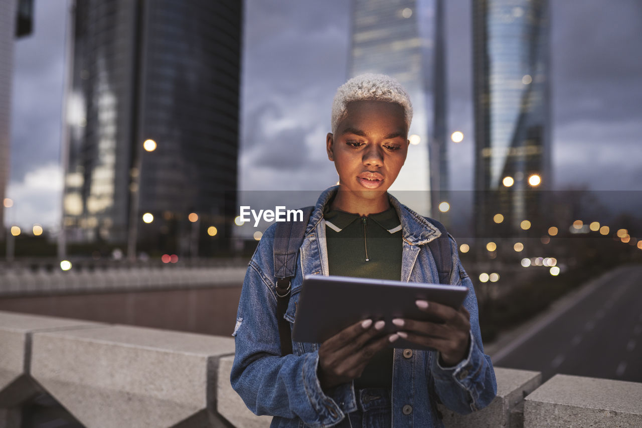 Woman with tablet pc standing on bridge in city at dusk