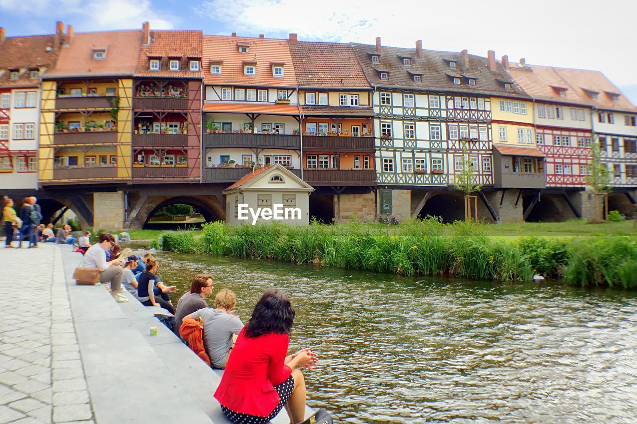 People sitting by river with buildings in background
