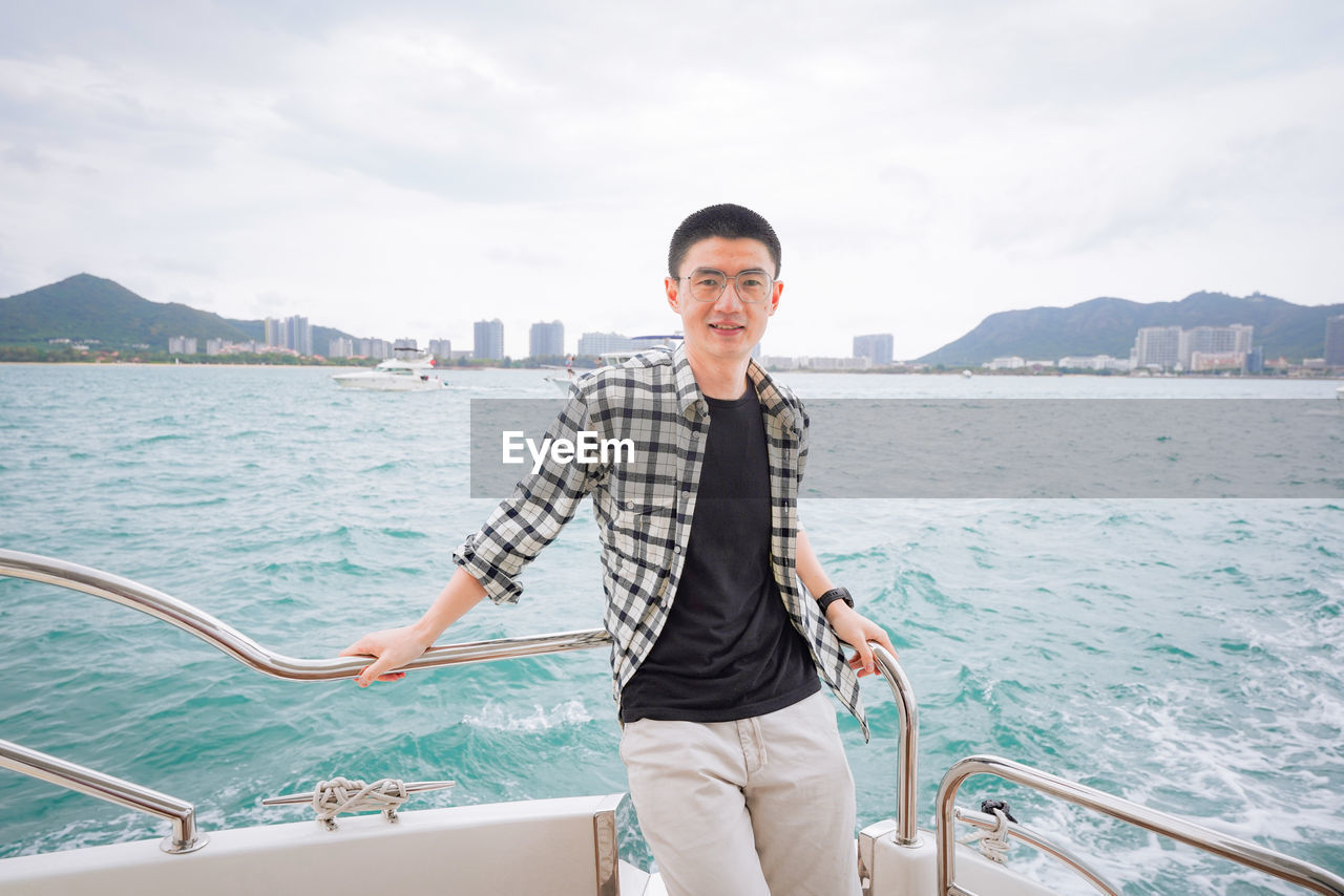 portrait of young man standing on boat in sea against sky