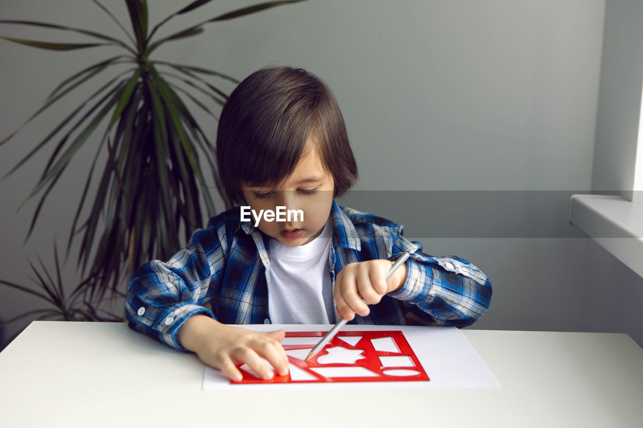 Boy child draws on paper with a ruler on a table sitting by the window