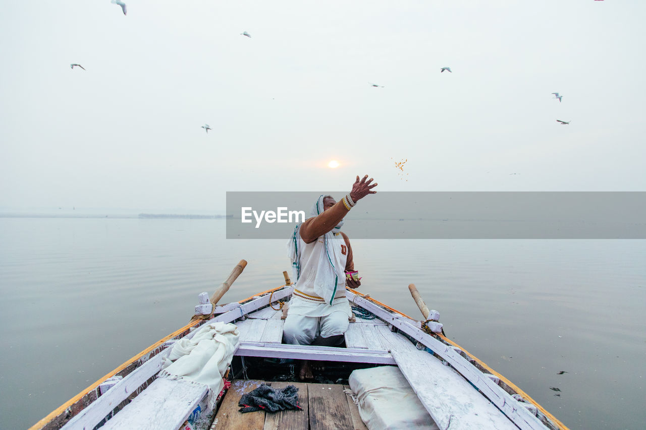 Varanasi, india - february, 2018: indian male in white clothes sitting in boat and throwing grains over river while feeding birds in early morning