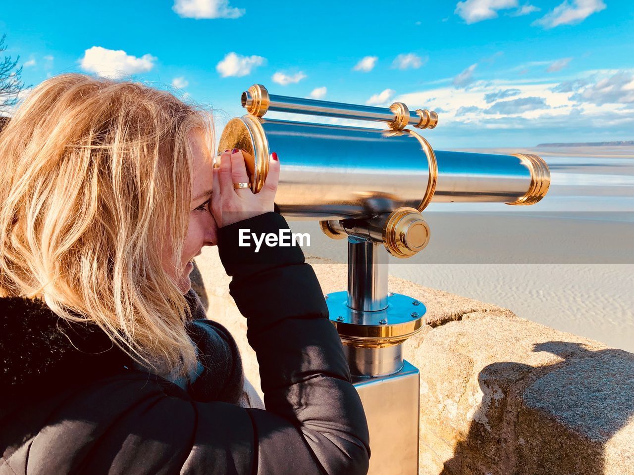 Side view of smiling woman looking through hand-held telescope at beach