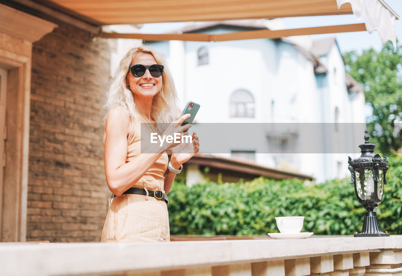 Adult elegant blonde woman in sunglasses and beige clothes using mobile phone at house backyard