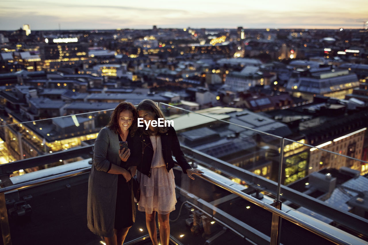 High angle view of smiling female professionals taking selfie while standing on terrace against illuminated cityscape