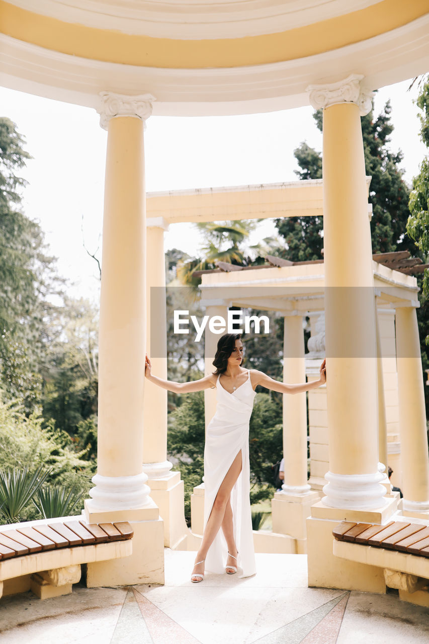 A beautiful brunette lady in an elegant wedding dress poses among the columns in the old city park