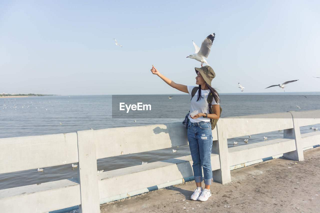 Full length of smiling woman feeding seagull standing against sea