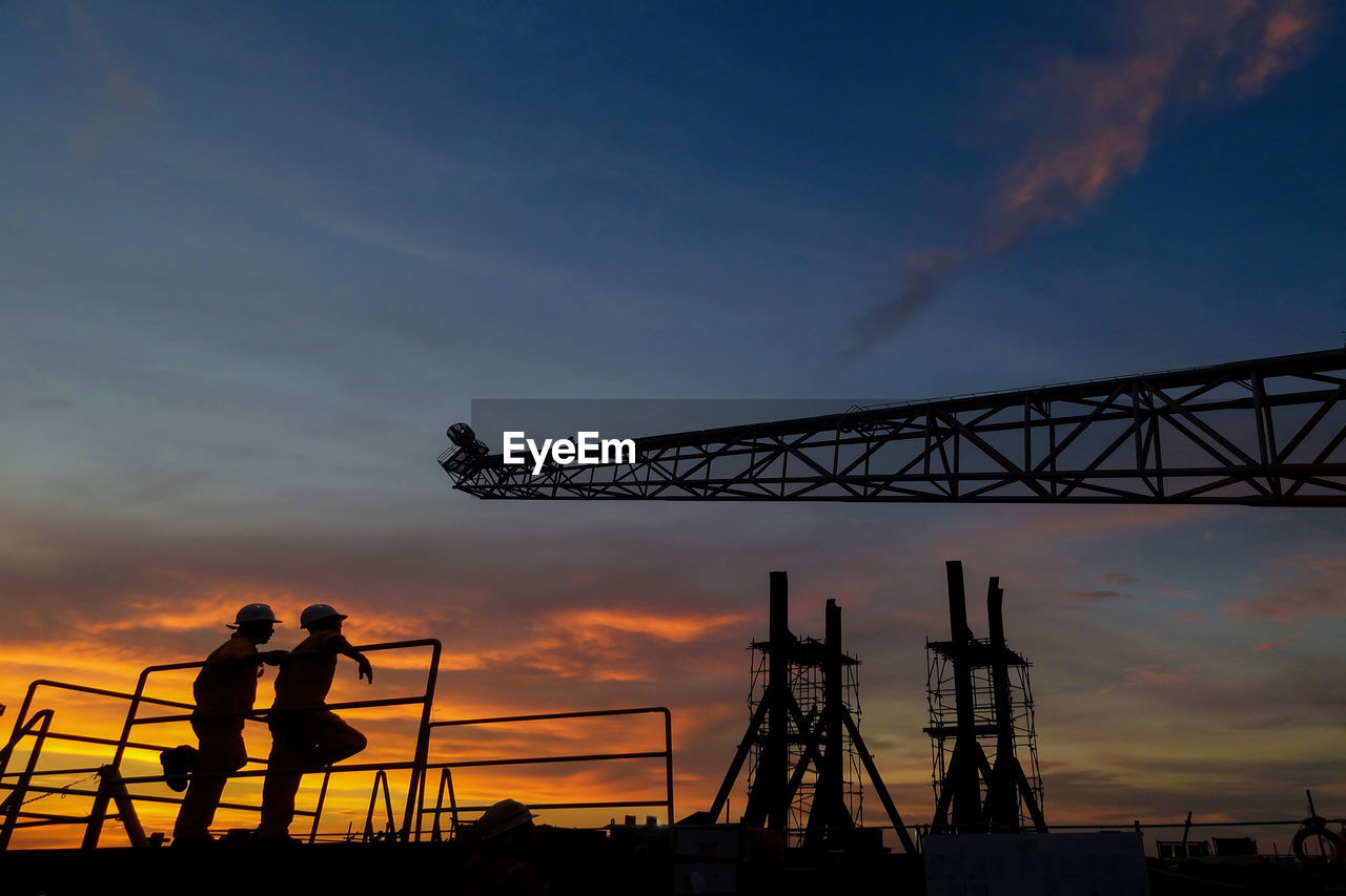 Workers leaning on railing by silhouette crane at construction site against sky during sunset