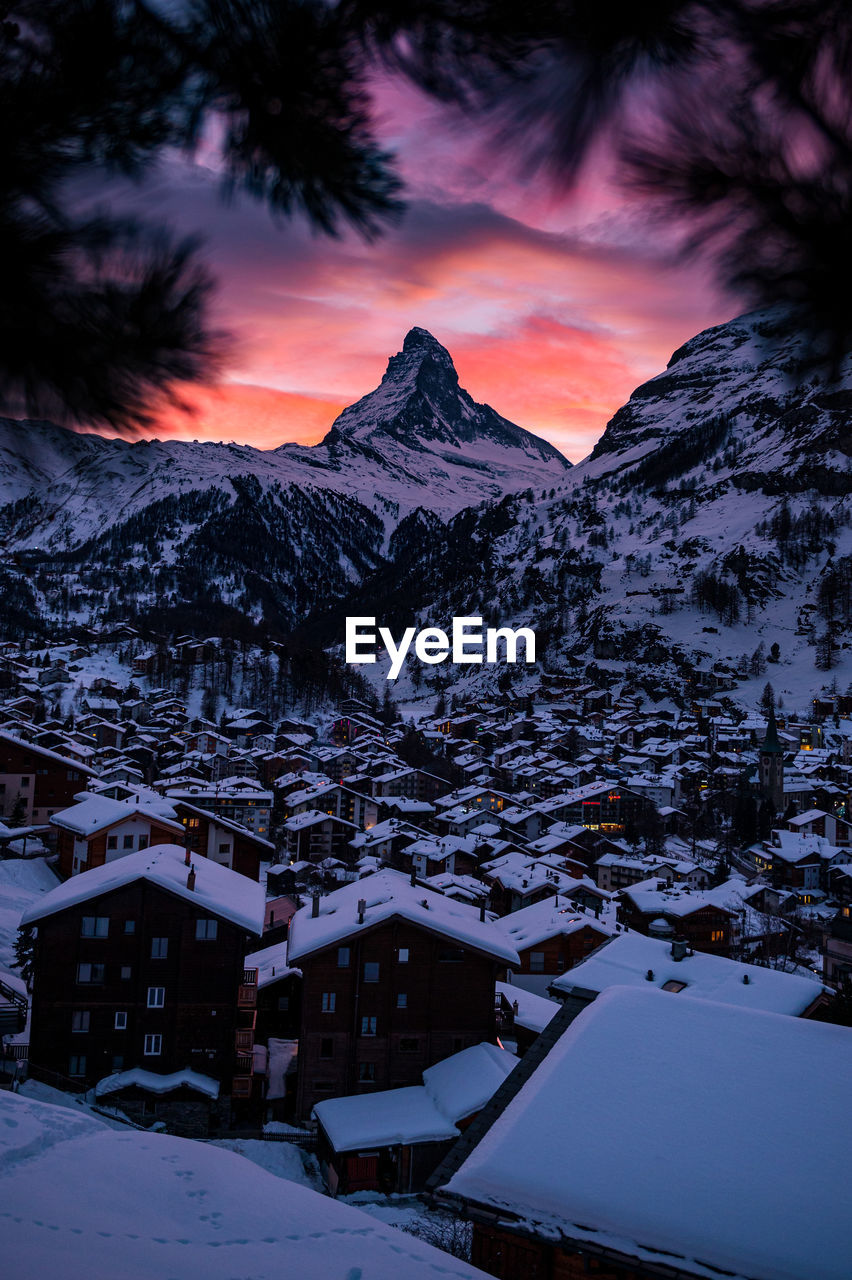 SNOW COVERED HOUSES AND MOUNTAINS AGAINST SKY AT SUNSET