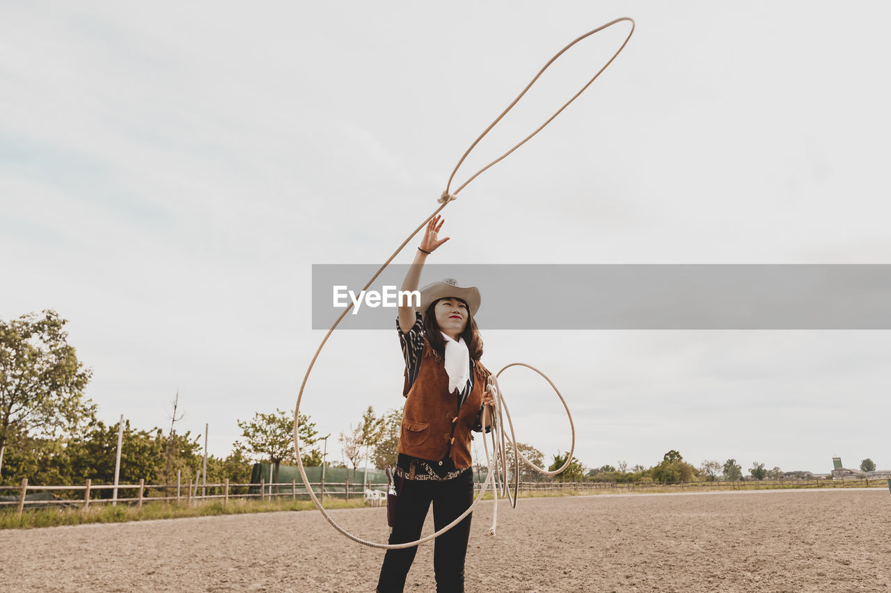 Cowgirl throwing lasso while standing at ranch against sky