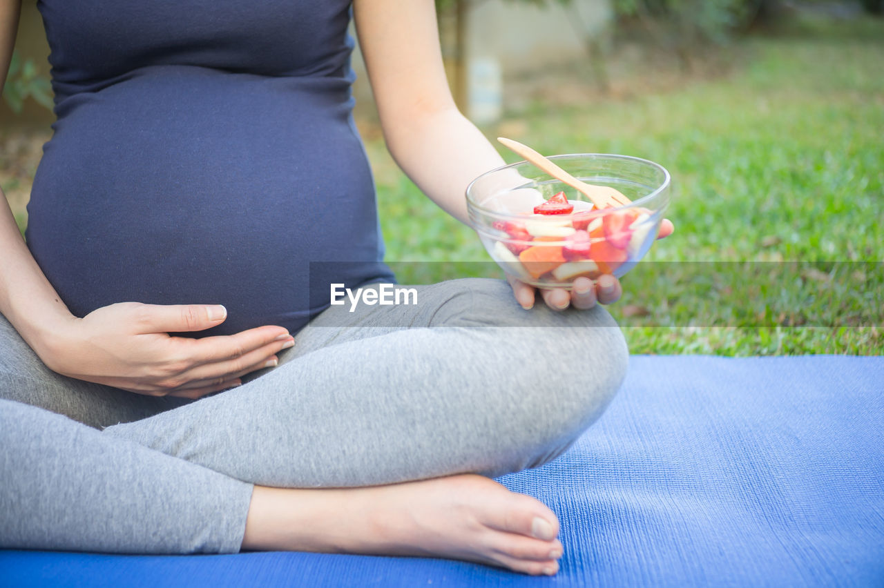 Low section of pregnant woman holding bowl with fruits while sitting on exercise mat