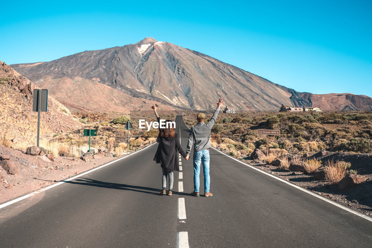 REAR VIEW OF MEN STANDING ON ROAD AGAINST SKY