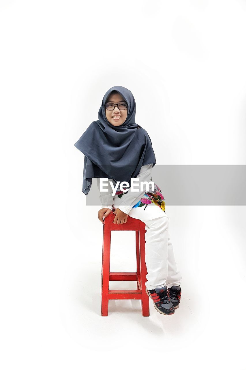 Portrait of smiling young woman wearing hijab sitting on stool against white background