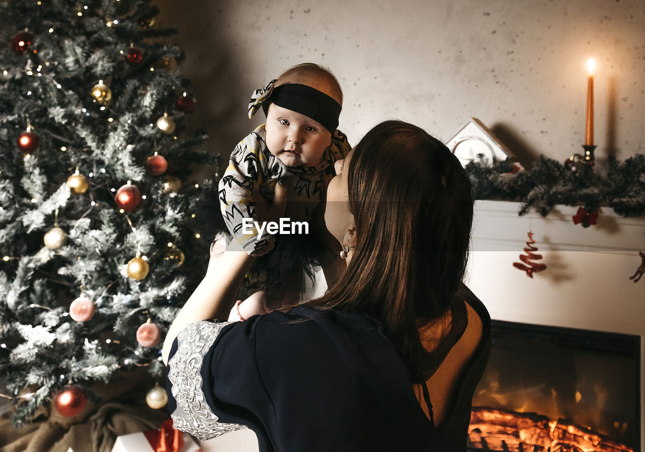 Woman holding baby girl by christmas tree at home