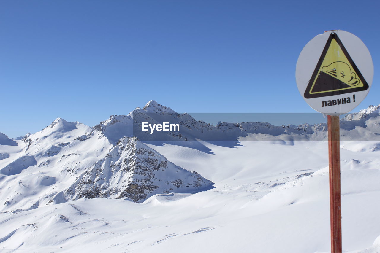 Warning sign by snow covered mountains against clear blue sky