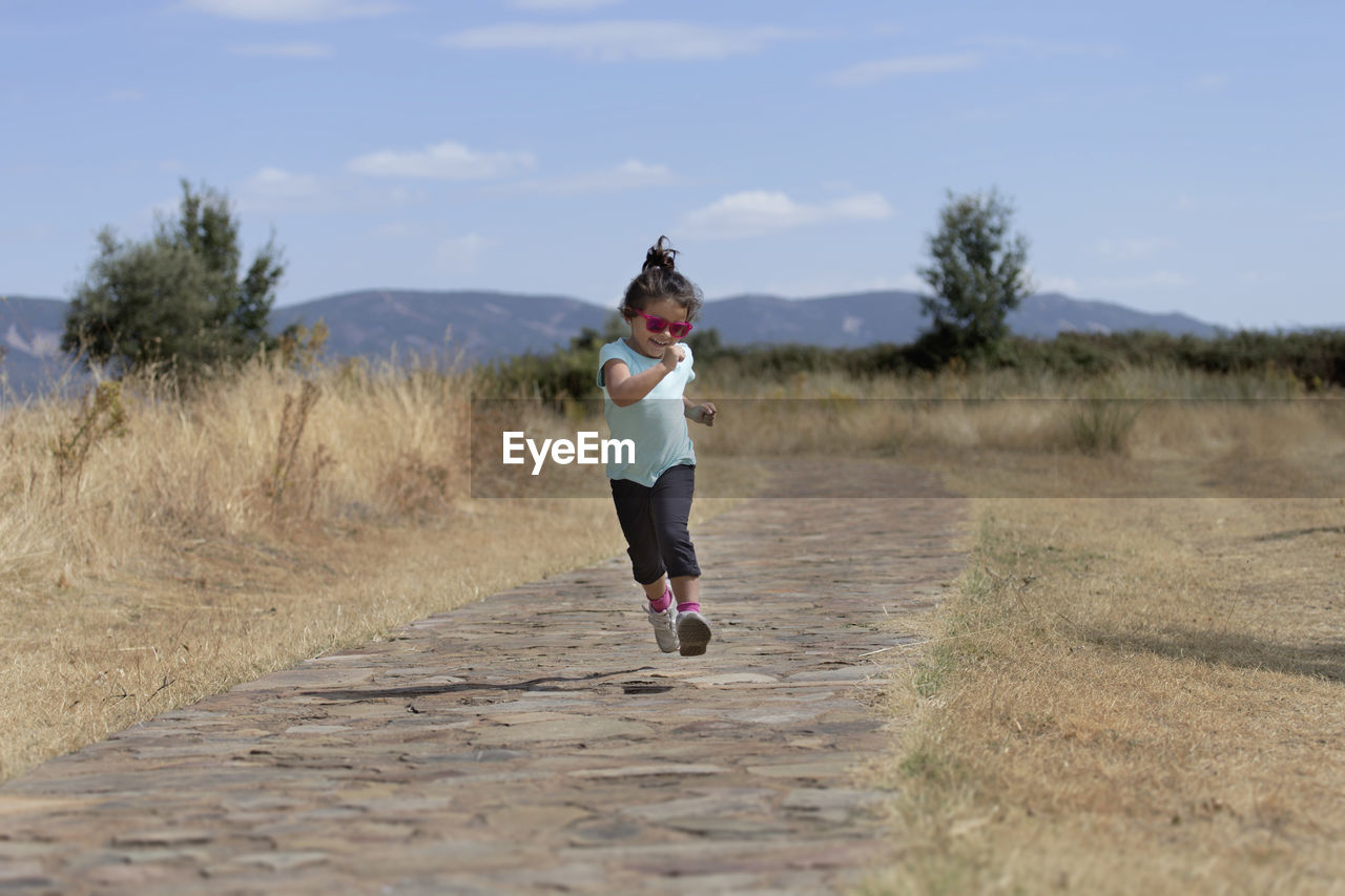 Full length of cheerful girl jumping on road amidst grassy field against sky