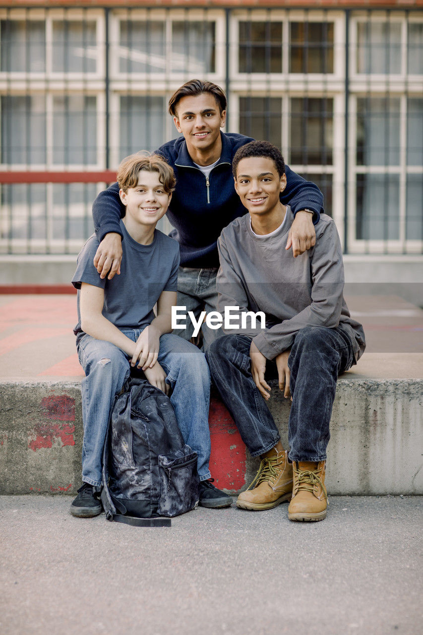 Portrait of smiling teenage student with arms around male friends sitting on concrete in high school campus