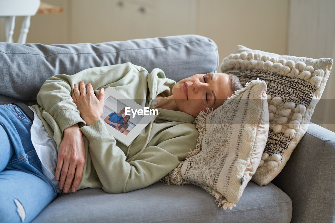 Sad mature woman holding family photo while lying with closed eyes on sofa at home
