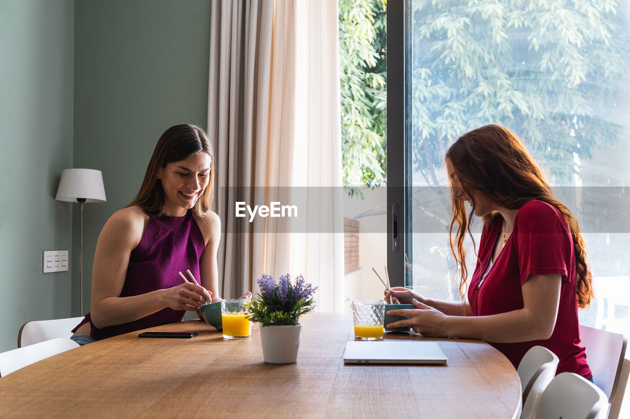 Happy young female friends in casual outfits having delicious lunch together and chatting while gathering at dining table in light apartment