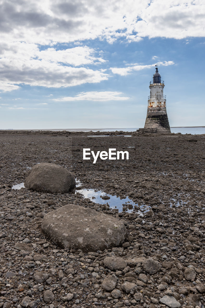 Rocks on beach with lighthouse by sea against sky